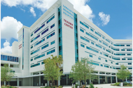 Large white hospital building with trees and cloudy sky.