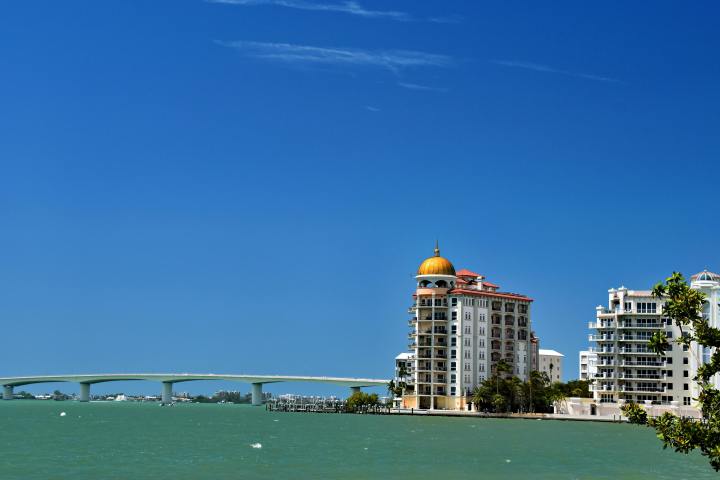 Coastal buildings with gold dome near a bridge over blue-green water under clear blue sky.