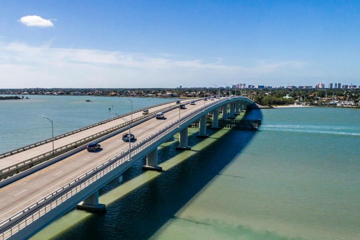 Cars crossing a long bridge over a blue-green river with buildings in the distance.